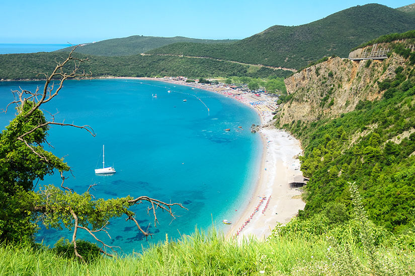 Weitläufiger Blick auf den Jaz Beach bei Budva mit türkisblauem Wasser und einem breiten Ufer, das aus Sand und Kies besteht. Im Hintergrund sieht man die geschwungene Bucht mit grüner Vegetation und Bereichen. Strände Montenegro Jaz Beach