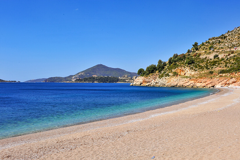 Ein breiter Sandstrand zieht sich in einer geschwungenen Bucht entlang, das Wasser wechselt direkt am Ufer von hellem Türkis zu tiefem Blau. Rechts rahmt eine felsige, teilweise bewachsene Hangseite die Küste ein, am Horizont liegt eine sanfte Bergsilhouette. Strände Montenegro Becici