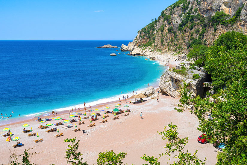 Top-Down-Blick auf eine kleine, gebogene Bucht mit leuchtend türkisfarbenem Wasser, das zum offenen Blau hin deutlich dunkler wird. Der helle Strandstreifen liegt direkt am grünen Hang, am Ufer erkennt man einzelne Schirme und Badegäste. Strände Montenegro Perazica Do