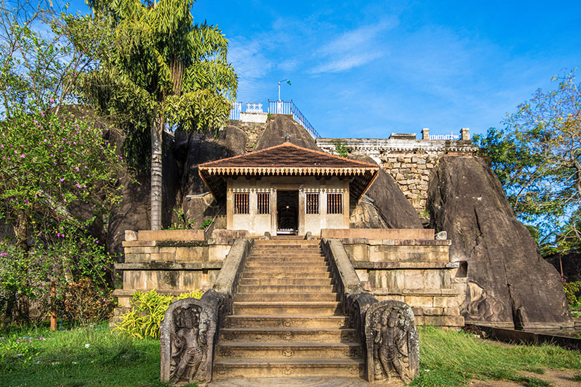 Ein historisches Tempelgebäude mit Schindeldach, das direkt in dunkle Felsformationen gebaut wurde, mit einer steilen Steintreppe im Vordergrund. Sri Lanka Isurumuniya Anuradhapura Sri Lanka Isurumuniya