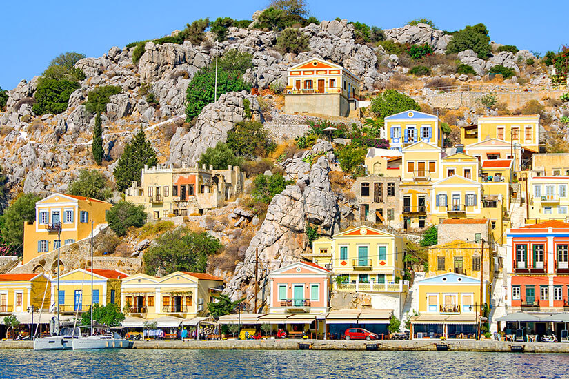 Bunte, renovierte Herren- und Kapitänshäuser in Pastellfarben ziehen sich am Hafen von Kalymnos den felsigen Hang hinauf. Im Vordergrund glitzert das Meer, dahinter rahmen graue Felsen und mediterranes Grün die Szenerie unter strahlend blauem Himmel. Kalymnos Griechenland Herren-und-Kapitanshauser