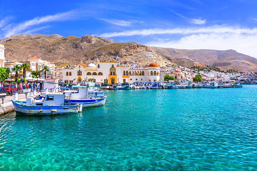 der Hafen von Pothia auf Kalymnos mit kristallklarem, türkisfarbenem Wasser im Vordergrund. Entlang der Promenade liegen kleine Boote, dahinter reihen sich helle Häuser und ein markantes Gebäude mit Kuppeln vor der kargen Bergkulisse. Über allem spannt sich ein intensiver blauer Himmel mit feinen Wolkenschleiern. Kalymnos Griechenland Pothia