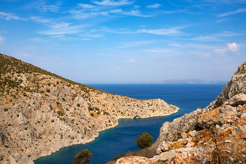 Der Blick zeigt die Vathi Bucht auf Kalymnos mit ruhigem Wasser und der geschützten, fjordartigen Einbuchtung zwischen den Hügeln. Am Ufer liegen kleine Boote und eine lockere Bebauung, die sich an die Küste schmiegt. Die karge, steinige Landschaft rahmt die Bucht unter klarem Himmel. Kalymnos Griechenland Vathi