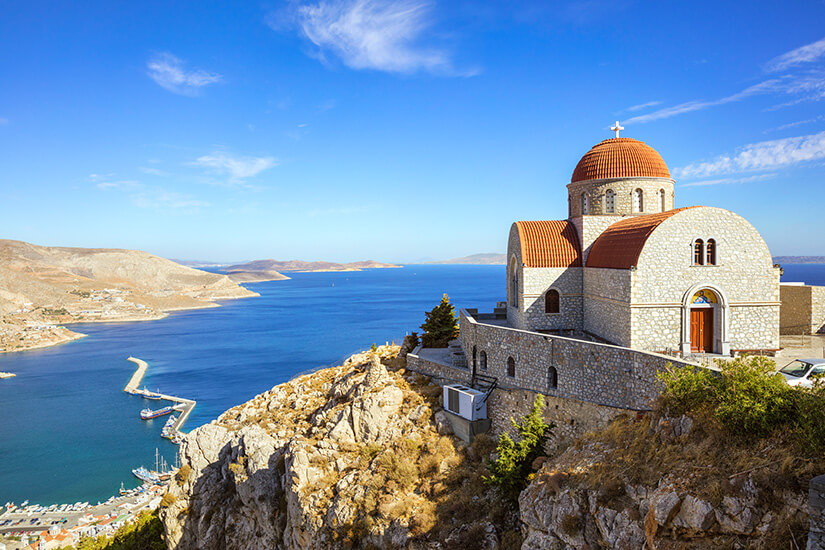 Das Kloster Agios Savvas thront oberhalb von Pothia auf Kalymnos und zieht sich terrassenförmig den Hang hinauf. Weiße Mauern, Treppen und Innenhöfe heben sich klar vom felsigen, braunen Berghang ab und wirken in der Sonne fast leuchtend. Im Hintergrund öffnet sich der Blick Richtung Stadt und Meer. Kalymnos Griechenland Agios Savvas