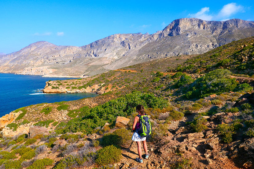 Eine Wanderin ist auf einem Pfad in der felsigen Landschaft von Kalymnos unterwegs. Um sie herum prägen karge Hänge, Steine und mediterranes Buschwerk das Bild, während der Blick in die Weite führt. Das klare Licht und der offene Himmel unterstreichen das Gefühl von Freiheit und Natur. Kalymnos Griechenland Wandern