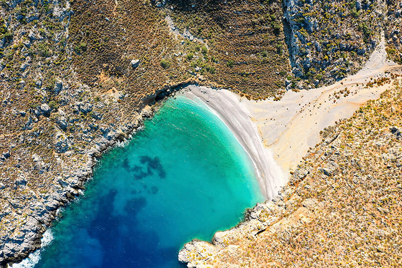 Geschwungene Bucht auf Kalymnos mit einem hellen Strand und kristallklarem, türkisfarbenem Wasser. Felsige Küstenlinien und karge Hänge rahmen die kleine Badebucht ein. Die Farbverläufe im Wasser zeigen flache Zonen am Ufer und tieferes Blau weiter draußen. Kalymnos Griechenland Strand