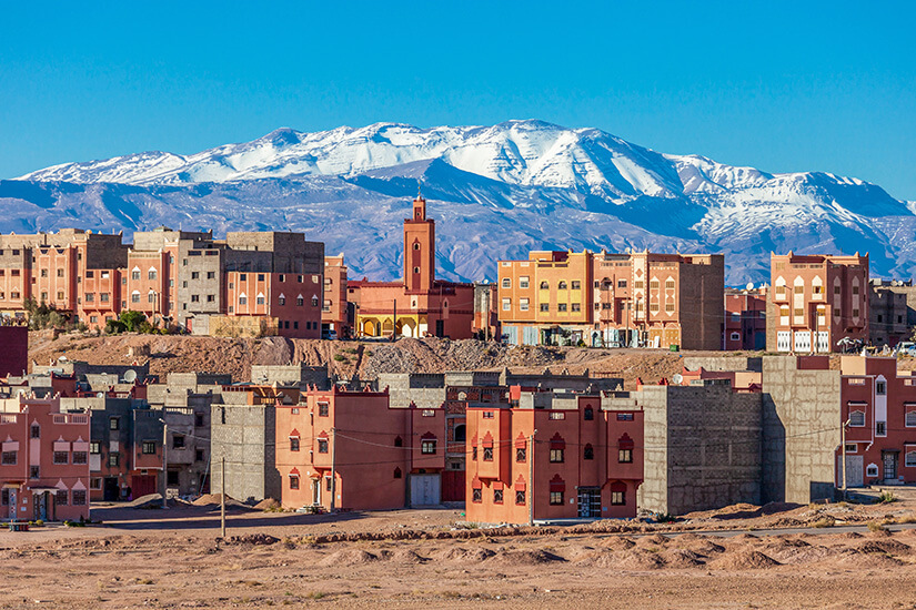 Eine Ansammlung flacher, erdfarbener Gebäude erstreckt sich in einer trockenen, sandigen Landschaft. In der Mitte ragt ein schlanker Turm zwischen den Häusern empor. Im Hintergrund erheben sich mächtige, schneebedeckte Berge unter einem klaren blauen Himmel. Ouarzazate Stadt