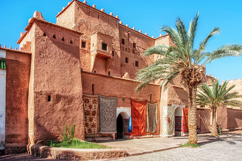 Eine aus Lehm errichtete Kasbah mit kunstvollen Verzierungen erhebt sich unter blauem Himmel. Vor den Torbögen hängen farbenfrohe Teppiche, daneben steht eine hohe Palme. Ouarzazate Taourirt Kasbah
