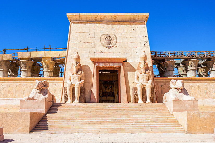 Ein monumentaler Tempelbau im altägyptischen Stil mit breiter Treppe und zentralem Eingang ist zu sehen. Beidseitig des Portals stehen große sitzende Pharaonenstatuen sowie Sphinx-Figuren, die den Aufgang flankieren. Reliefs und Säulen mit hieroglyphenähnlichen Verzierungen schmücken die Fassade unter klarem blauem Himmel. Ouarzazate Filmstudio