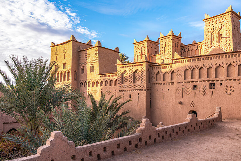 Eine große Kasbah aus rötlichem Lehm mit verzierten Fassaden und eckigen Türmen dominiert die Szene. Palmen wachsen im Innenhofbereich, während eine niedrige Mauer mit dekorativen Zinnen im Vordergrund entlangführt. Ouarzazate Amridil