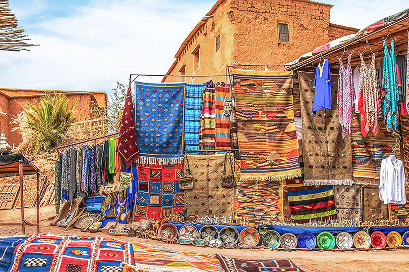 Ein Marktstand im Freien bietet bunte Teppiche, Decken und Tücher mit geometrischen Mustern an. Vor dem Stand liegen bemalte Keramikteller und Schalen, daneben hängen Kleidungsstücke und Taschen. Im Hintergrund stehen ockerfarbene Lehmgebäude. Ouarzazate Markt