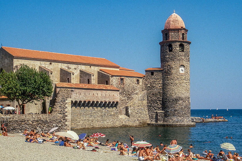 Sommerliche Szene am Kieselstrand Plage de Boramar in Collioure mit zahlreichen Badegästen unter Sonnenschirmen und der markanten Kirche Notre-Dame-des-Anges mit ihrem runden Glockenturm direkt am blauen Meer. Collioure Frankreich Plage de Boramar