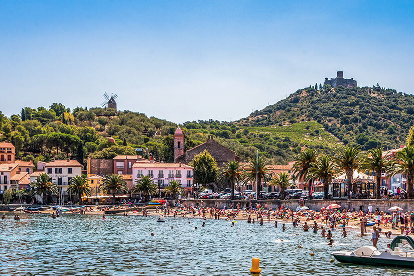 Blick über das türkisfarbene Wasser am Plage du Port d’Avall in Collioure mit Badegästen im Meer, bunten Tretbooten am flachen Sandstrand und der historischen Kulisse der Stadt mit Windmühlen auf den grünen Hügeln im Hintergrund. Collioure Frankreich Plage du Port d'Arvall
