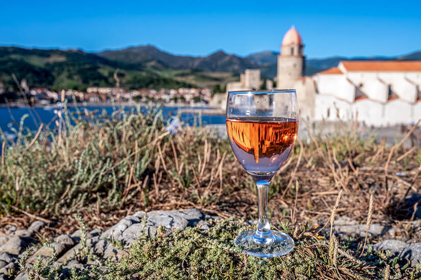 Ein Glas gekühlter Roséwein auf einem Stein im Vordergrund, in dem sich die Kirche Notre-Dame-des-Anges spiegelt, mit dem Hafen von Collioure und den Pyrenäen im unscharfen Hintergrund. Collioure Frankreich Wein