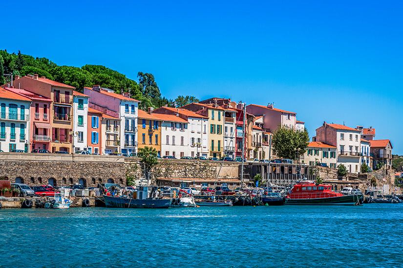 Blick vom blauen Meer auf die Hafenpromenade von Port-Vendres mit farbenfrohen Häuserfassaden, Fischerbooten und einem auffälligen roten Rettungskreuzer vor grünen Bäumen unter klarem Himmel. Collioure Frankreich Port Vendres