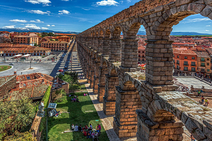 Von einem erhöhten Standpunkt aus fällt der Blick über die Dächer Saragossas, während im Vordergrund die mächtigen Steinbögen eines römischen Aquädukts die Szenerie dominieren. Die warmen Erdtöne der Altstadt kontrastieren mit dem satten Grün der Vegetation am Fuße des Bauwerks. Im Hintergrund erstreckt sich die Stadt bis zu den sanften Hügeln am Horizont. Städte Spanien Saragossa