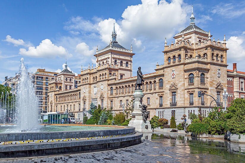 Die prachtvolle Academia de Caballería erhebt sich wie ein Märchenschloss mitten in Valladolid, mit ihren neogotischen Türmen und der reich verzierten Fassade. Vor dem Gebäude plätschert ein großer Springbrunnen, dessen Wasserfontäne das Sonnenlicht einfängt. Eine Denkmalstatue vervollständigt diese Szene spanischer Grandezza. Städte Spanien Valladolid