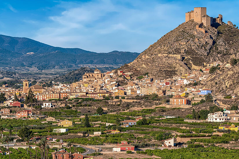Wie ein Adlerhorst thront eine mittelalterliche Burg auf dem kahlen Felsgipfel über einem typisch spanischen Bergdorf mit seinen ockerfarbenen Häusern und der markanten Kirchturmspitze. Terrassierte Obstgärten und grüne Felder umgeben die Siedlung wie ein fruchtbarer Gürtel. Städte Spanien Murcia