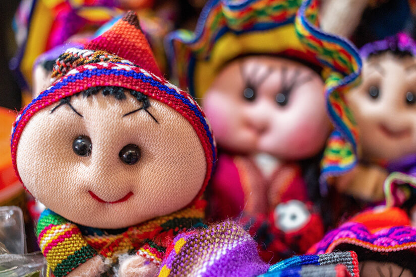 Mehrere kleine Stoffpuppen mit runden Gesichtern und großen Augen sitzen dicht nebeneinander. Sie tragen farbenfrohe, traditionell wirkende Kleidung mit gewebten Mustern. Souvenirs Chile Puppen