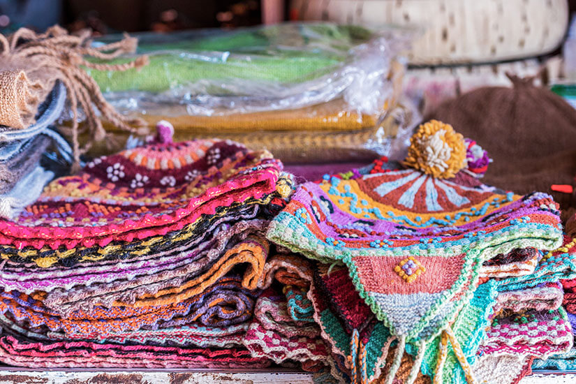 Mehrere farbenfrohe Strickwaren liegen ordentlich gestapelt auf einem Tisch. Die Mützen und Textilien zeigen verspielte Muster, Fransen und kleine Bommel. Souvenirs Chile Chullo