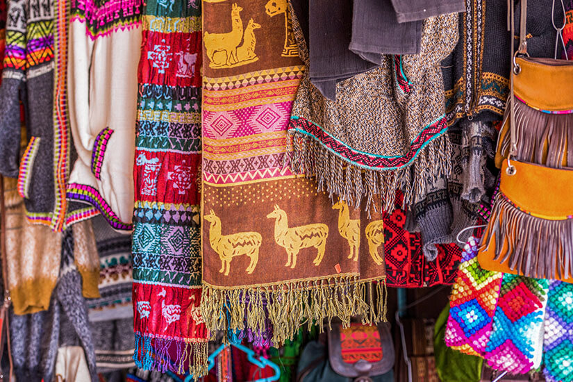 Mehrere bunte Schals und Textilien hängen dicht nebeneinander an einem Verkaufsstand. Die Stoffe zeigen auffällige Muster, Fransen und tierische Motive wie Lamas. Souvenirs Chile Schals