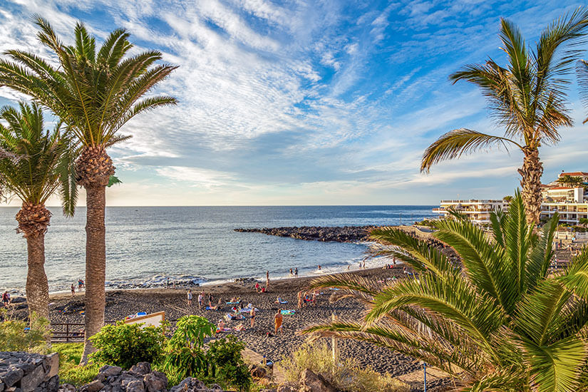 Ein Strand mit dunklem Vulkansand an der Küste von Teneriffa. Grüne Palmen rahmen das Bild. Puerto de Santiago Spanien Wetter