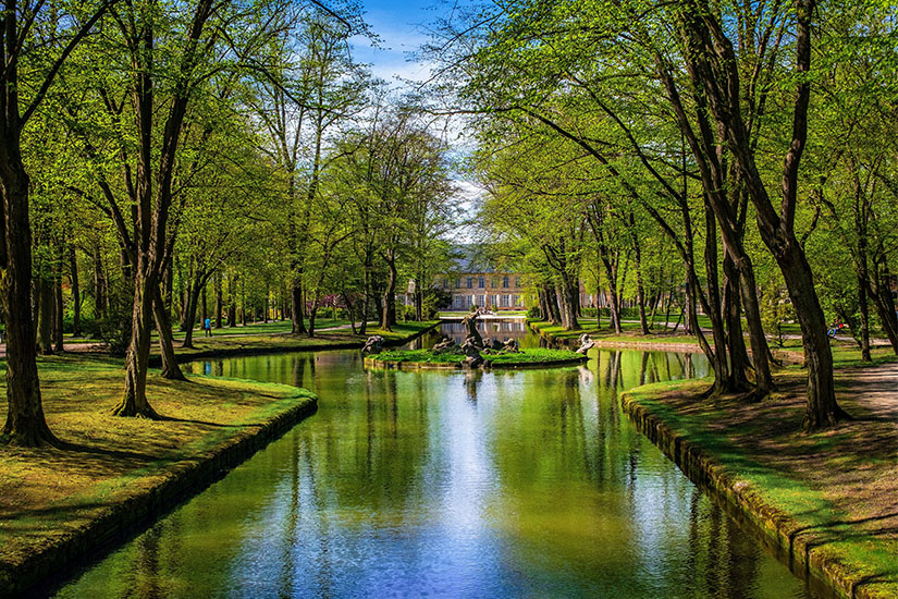 Ein ruhiger Kanal führt durch eine von alten Laubbäumen gesäumte Parkanlage auf ein elegantes Gebäude zu. Das frische Grün spiegelt sich malerisch im Wasser. Die Anlage verbindet französische Gartenkunst mit oberfränkischer Idylle. Bayreuth Deutschland Schlosspark