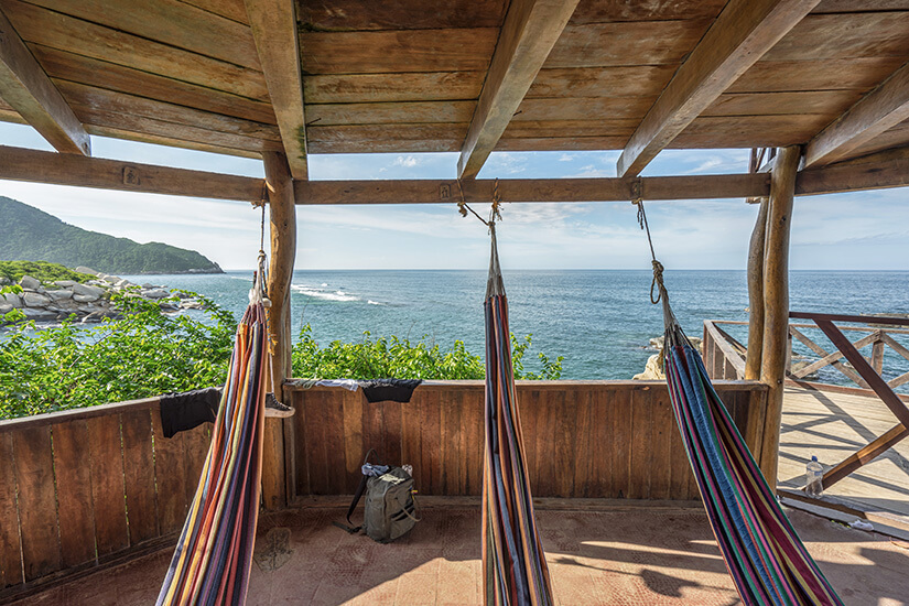 Bunte Hängematten unter einem Holzgebälk mit direktem Blick auf das türkisfarbene Meer am Cabo San Juan. Tayrona Nationalpark Hängematten Cabo San Juan