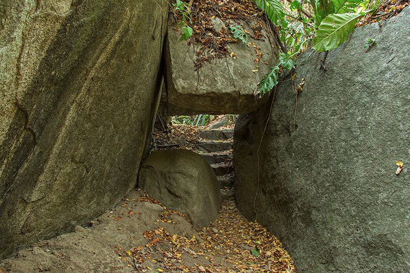 Ein schmaler Pfad führt durch eine enge Passage zwischen massiven Felsblöcken im dichten Küstenwald. Tayrona Nationalpark Sendero de las 9 Piedras