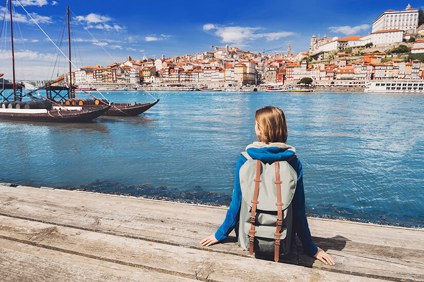 Eine Urlauberin mit Rucksack sitzt am Ufer und blickt über einen breiten Fluss auf eine dicht bebaute Altstadt. Im Wasser liegen traditionelle Holzboote vor der Kulisse farbenfroher Häuser am Hang. Packliste Portugal Hafen
