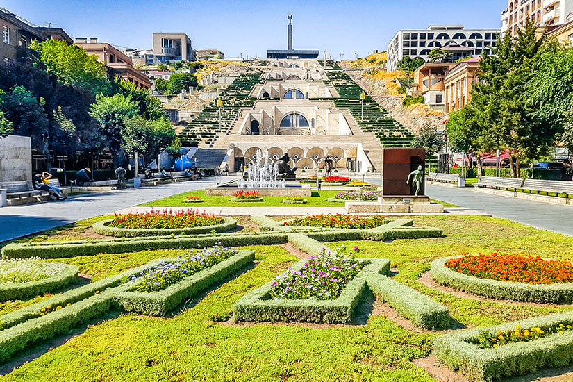 Eine breite Treppenanlage mit begrünten Terrassen und Springbrunnen führt einen Hügel hinauf. Im Vordergrund sind kunstvoll angelegte Blumenbeete und gepflegte Rasenflächen zu sehen. Oben auf dem Hügel steht ein Denkmal, umgeben von modernen und historischen Gebäuden. Armenien Fruehling