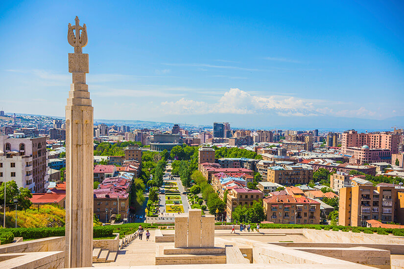 Von einer erhöhten Terrasse blickt man über eine weitläufige Stadt mit dicht bebauten Wohn- und Geschäftsvierteln. Im Vordergrund steht eine hohe Steinsäule mit einem ornamentalen Symbol, dahinter führen Treppen und Wege hinunter in einen grünen Park. Am Horizont erhebt sich ein schneebedecktes Gebirge unter klarem Himmel. Armenien Jerewan
