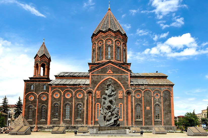 Eine große Kirche aus dunklem Stein mit rötlichen Verzierungen und zwei markanten Türmen steht unter blauem Himmel. Vor dem Gebäude befindet sich ein modernes Denkmal aus Stein mit mehreren Figuren. Die Fassade ist mit Rundbögen und dekorativen Elementen im traditionellen Stil gestaltet. Armenien Gjumri