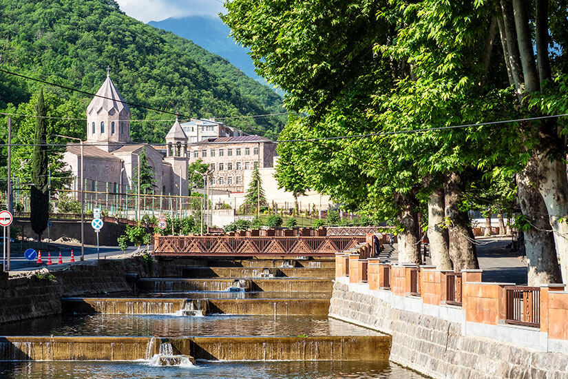 Ein kanalartig gefasster Fluss mit kleinen Kaskaden fließt durch eine grüne Stadtlandschaft. Entlang des Ufers stehen Bäume und eine Promenade mit Geländern, im Hintergrund erheben sich bewaldete Berge. Zwischen den Hügeln sind kirchliche Gebäude mit spitzen Dächern zu erkennen. Armenien Kapan