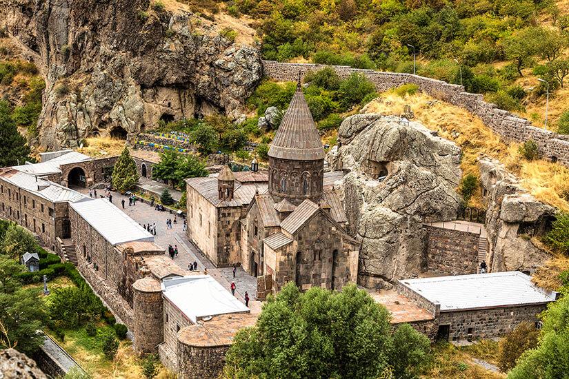 Ein historisches Kloster aus hellem Stein liegt eingebettet in eine felsige Schlucht. Die Anlage ist von Mauern umgeben und wird von steilen Felswänden überragt, in die teilweise Höhlen eingelassen sind. Mehrere Besucher bewegen sich im Innenhof zwischen den Gebäuden mit konischem Kirchturm. Armenien Geghard