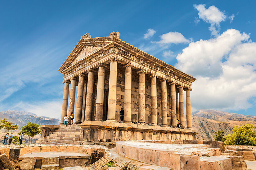 Ein antiker Tempel mit zahlreichen Säulen steht auf einem erhöhten Plateau vor bergiger Kulisse. Das Bauwerk aus hellem Stein besitzt ein dreieckiges Giebeldach im klassischen Stil. Einige Besucher befinden sich auf den Stufen und zwischen den Säulen. Garni Tempel