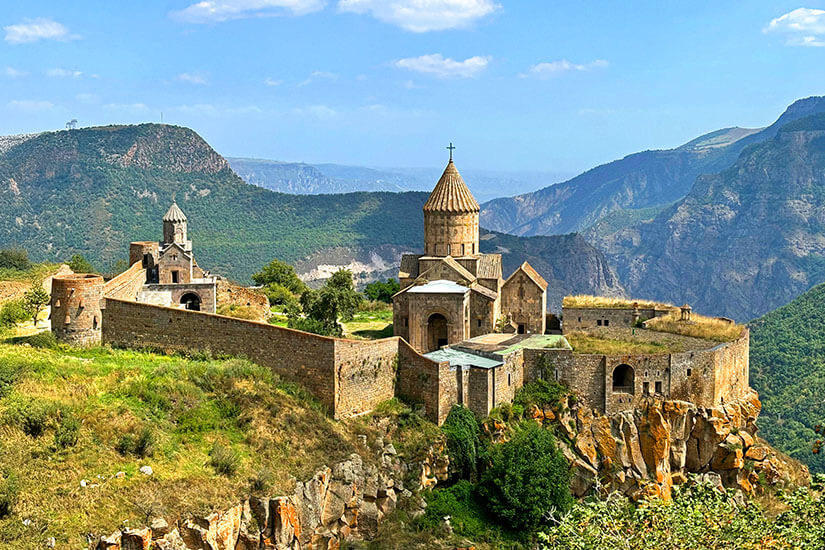 Ein altes Klosterensemble aus Stein thront auf einer steilen Felskante über einer tiefen Schlucht. Mehrere Gebäude mit konischen Kirchtürmen sind von einer Mauer umgeben und liegen inmitten grüner Hügel. Im Hintergrund erstrecken sich beeindruckende Berglandschaften unter blauem Himmel. Armenien Tatev