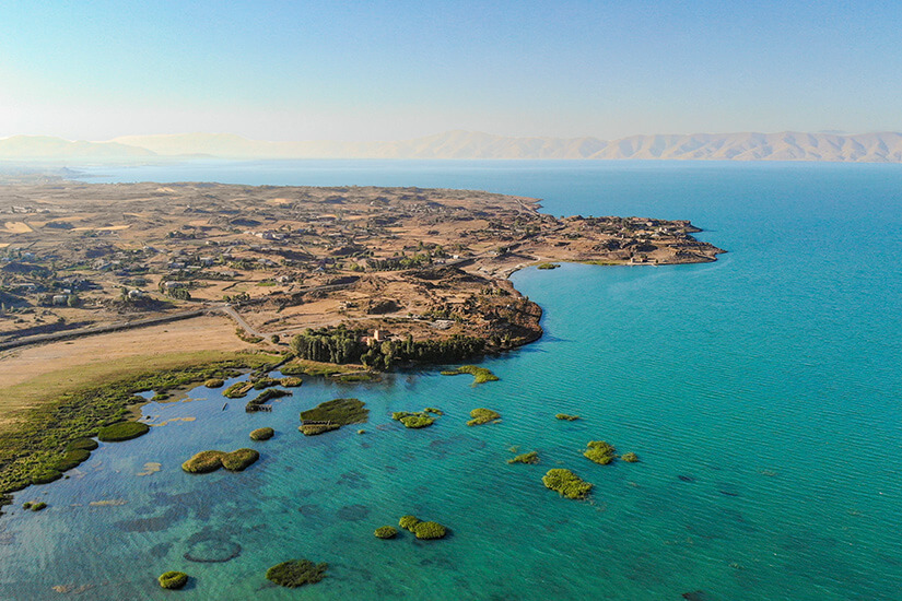 Eine weitläufige Küstenlandschaft mit türkisfarbenem Wasser breitet sich unter klarem Himmel aus. Kleine bewachsene Inseln oder Schilfflächen ragen aus dem flachen Uferbereich hervor, während dahinter trockene Felder und verstreute Gebäude liegen. In der Ferne zeichnen sich sanfte Bergketten am Horizont ab. Sewan See