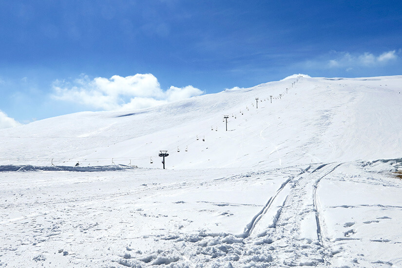 Eine weitläufige, schneebedeckte Berglandschaft breitet sich unter blauem Himmel aus. Entlang des Hanges verläuft eine Reihe von Skiliften mit Masten und Sesseln. Im Vordergrund sind Spuren im frischen Schnee zu erkennen. Armenien Skigebiet