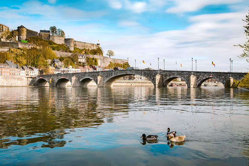 Eine historische Steinbrücke mit mehreren Bögen überspannt einen ruhigen Fluss, auf dem Enten schwimmen, während im Hintergrund eine mächtige Zitadelle auf einem Felsplateau thront. Staedte Belgien Namur