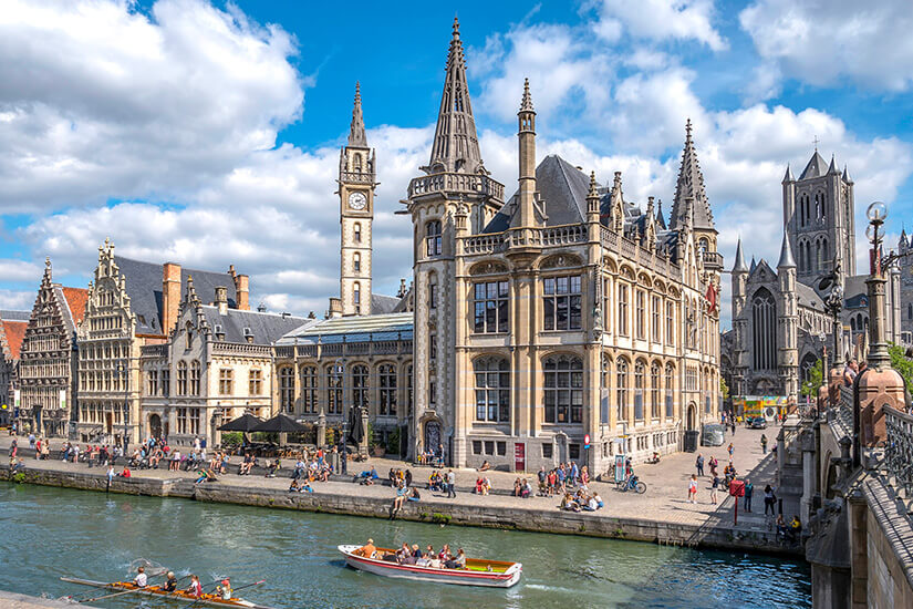 Prächtige gotische Gildehäuser und der imposante Belfried säumen die belebte Uferpromenade eines Kanals, auf dem Ausflugsboote gleiten, während Menschen das geschäftige Treiben genießen. Staedte Belgien Gent