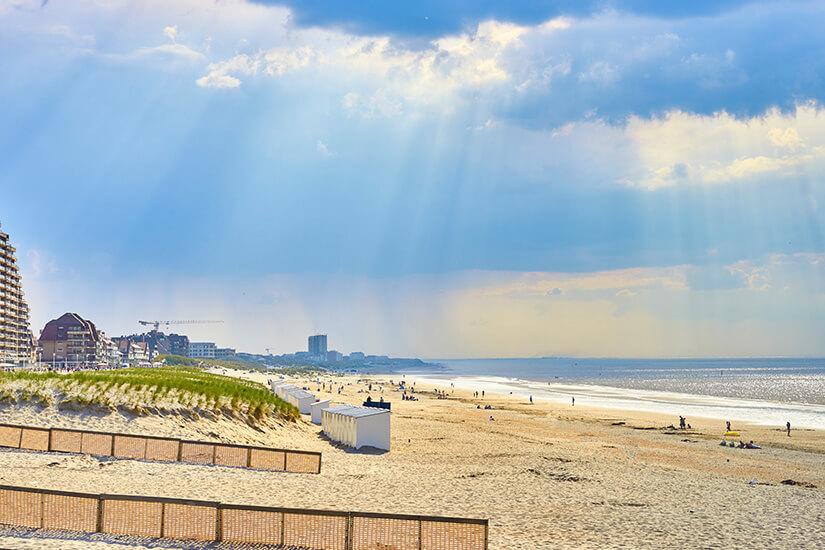 Dramatische Sonnenstrahlen brechen durch die Wolkendecke über einem breiten Sandstrand, auf dem verstreute Badegäste und die typischen weißen Kabinen zu sehen sind. Staedte Belgien Nieuwpoort