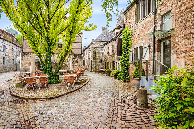 Eine geschwungene Gasse schlängelt sich zwischen ehrwürdigen Natursteinhäusern hindurch, während unter einem schattenspendenden Baum Caféstühle zum Verweilen einladen und Efeu die historischen Fassaden hinaufklettert. Staedte Belgien Durbuy