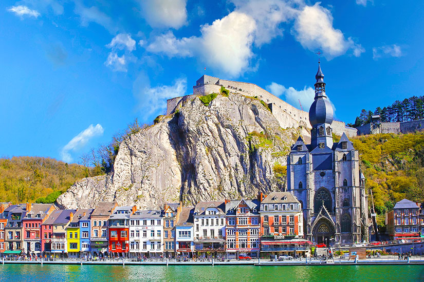 Eine mächtige Zitadelle thront auf einem steilen Kalksteinfelsen über bunten Häuserzeilen am Flussufer, während die gotische Stiftskirche Notre-Dame mit ihrem markanten zwiebelförmigen Turm das Stadtbild prägt. Staedte Belgien Dinant