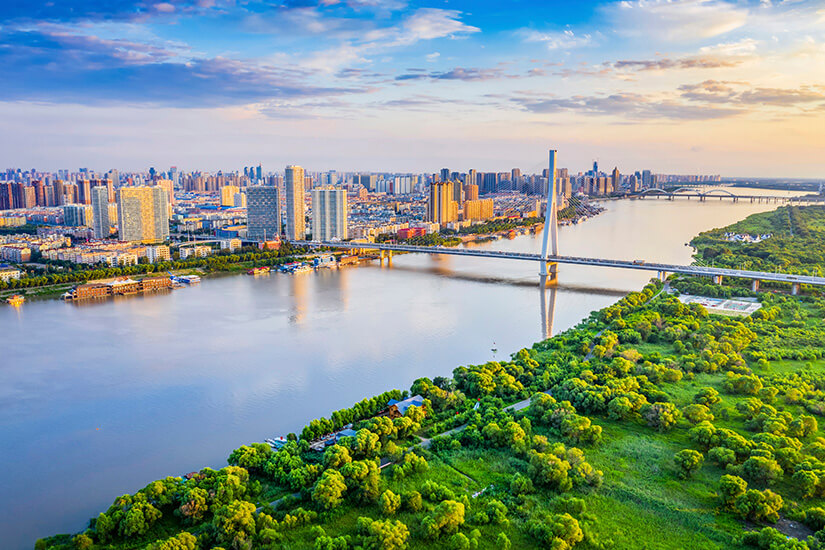 Eine moderne Großstadt erstreckt sich entlang eines breiten Flusses. Mehrere Brücken, darunter eine markante Schrägseilbrücke, verbinden die beiden Ufer mit Hochhäusern und Wohnkomplexen. Im Vordergrund liegt eine grüne Parklandschaft, während die Sonne die Skyline in warmes Licht taucht. Harbin Stadt