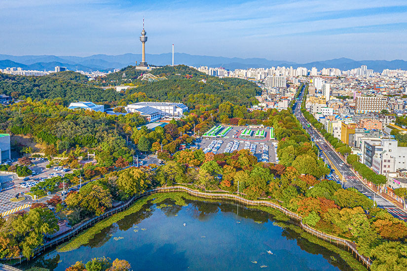 Aus der Vogelperspektive zeigt sich der Duryu Park als grüne Oase inmitten des Stadtgebiets, mit einem Teich im Vordergrund und dem 83 Tower auf der Anhöhe dahinter. Die herbstliche Verfärbung der Bäume setzt farbliche Akzente zwischen den umliegenden Straßenzügen. Die Aufnahme verdeutlicht, wie viel Grünfläche Daegu trotz seiner Größe bietet. Daegu Südkorea Vogelperspektive
