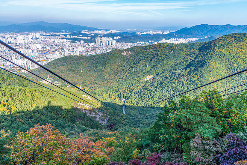 Die Apsan-Seilbahn gleitet über dicht bewaldete Hänge, während sich im Hintergrund das Stadtpanorama von Daegu ausbreitet. Die herbstliche Laubfärbung mischt Grün, Orange und Rot entlang der Bergkämme. Von oben wird sichtbar, wie nah Natur und Großstadt hier beieinanderliegen. Daegu Südkorea Apsan
