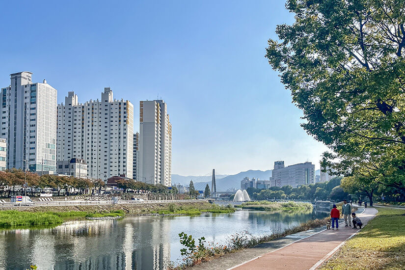 Ein gepflegter Uferweg führt entlang eines ruhigen Flusses, dahinter erheben sich Wohnhochhäuser und Berge im Dunst. Ein Springbrunnen sorgt für Bewegung auf dem Wasser, Spaziergänger nutzen den Weg am Nachmittag. Das Bild zeigt den grünen, alltagsnahen Charakter der Stadt. Daegu Südkorea Sincheon