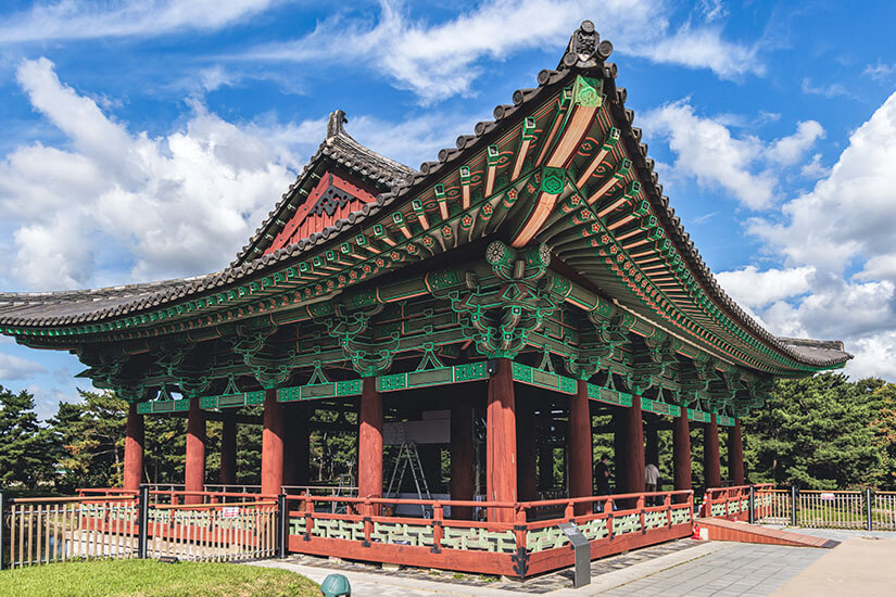 Ein kunstvoll verzierter koreanischer Pavillon mit geschwungenem Dach und detailreichen Holzarbeiten in Grün und Rot steht vor einem weiten Wolkenhimmel. Die Bauweise zeigt klassische Merkmale der Joseon-Architektur. Der offene Aufbau gibt den Blick in alle Richtungen frei. Daegu Südkorea Gyeongju