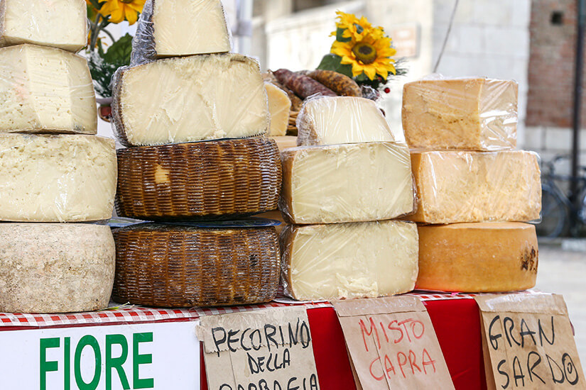 Mehrere große Käselaibe und -stücke sind auf einem Marktstand übereinandergestapelt. Beschriftete Schilder weisen auf Sorten wie Pecorino, Mischkäse aus Ziegenmilch und andere regionale Spezialitäten hin. Sonnenblumen und Wurstwaren dekorieren den Stand im Hintergrund. Souvenirs Sardinien Kaese
